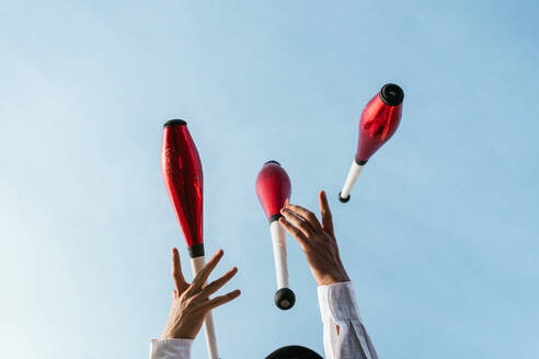 From below of crop anonymous circus artist performing trick with juggling clubs against blue sky - ADSF29140