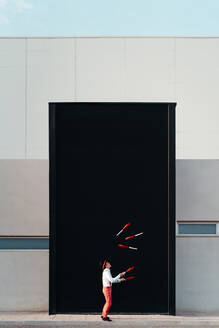 Seitenansicht eines nicht erkennbaren männlichen Zirkusjongleurs, der einen Trick mit Jonglierkeulen vor einer hohen schwarzen Wand eines modernen Gebäudes vorführt - ADSF29141