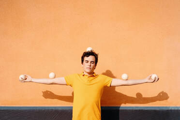 Tranquil immobile young male in bright yellow shirt with juggling balls on head and outstretched arms standing with eyes closed against orange wall - ADSF29150
