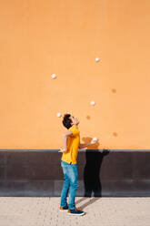 Full body side view of young male performing trick with juggling balls while standing on pavement near bright orange wall - ADSF29151