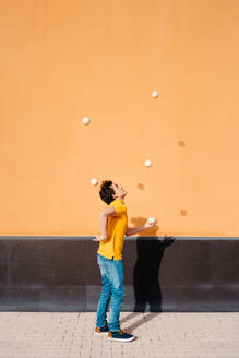 Ganzkörper-Seitenansicht eines jungen Mannes, der einen Trick mit Jonglierbällen vorführt, während er auf dem Bürgersteig in der Nähe einer leuchtend orangefarbenen Wand steht - ADSF29151