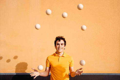 Happy young talented male performing trick with juggling balls while standing looking at camera on pavement near bright orange wall - ADSF29152