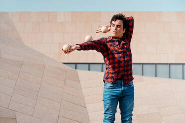 Skilled young male in checkered shirt performing trick with juggling balls while standing against contemporary concrete structure on urban street - ADSF29154