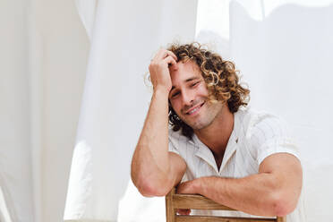 Delighted handsome male with curly hair sitting on chair and leaning on hand while looking at camera on background of white curtains - ADSF29166