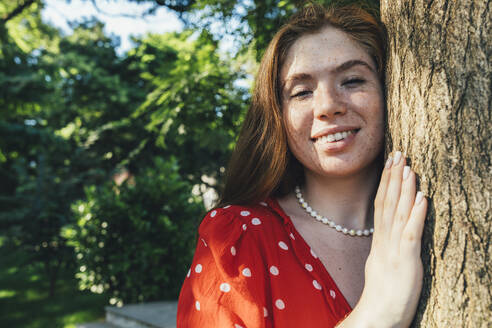 Lächelnde Frau, die an einem Baum in einem öffentlichen Park steht - OYF00414