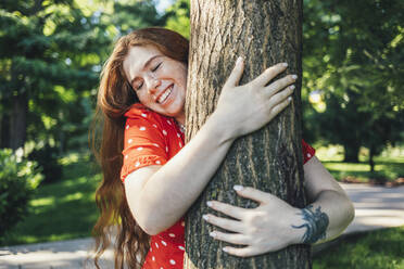 Glückliche junge Frau umarmt Baum mit geschlossenen Augen im Park - OYF00417