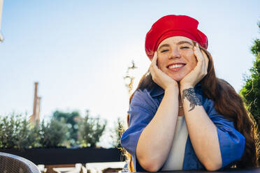 Smiling young woman with hands on chin at sidewalk cafe - OYF00425