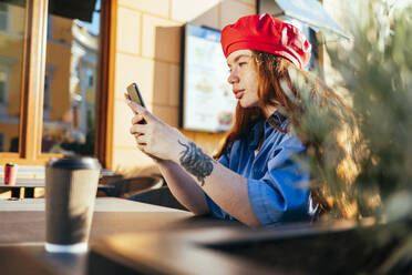 Junge Frau mit Baskenmütze benutzt ihr Smartphone in einem Straßencafé - OYF00435