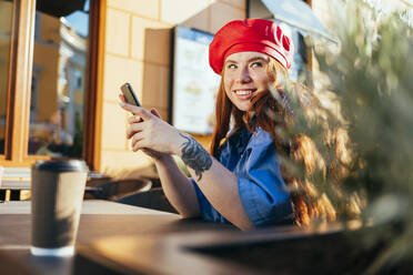 Smiling woman with mobile phone at cafe in sunlight - OYF00436