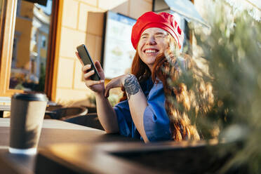 Junge Frau lacht, während sie ihr Smartphone in einem Straßencafé hält - OYF00438
