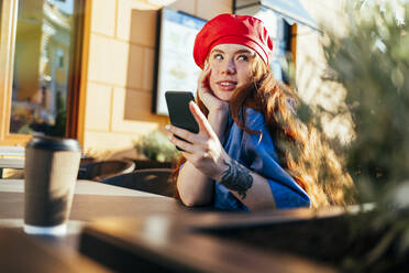 Young woman in beret hat day dreaming while holding mobile phone at sidewalk cafe - OYF00441