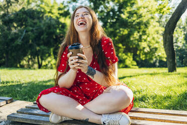 Glückliche rothaarige Frau mit Kaffeetasse sitzend im Park - OYF00476
