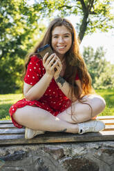 Young woman holding disposable cup while sitting at park - OYF00479