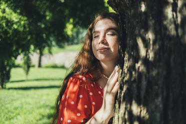Rothaarige Frau mit geschlossenem Auge umarmt Baum im Park - OYF00484