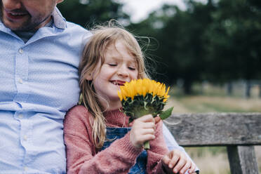 Lächelndes Mädchen, das eine Blume hält und mit seinem Vater auf einer Bank sitzt - ASGF01291