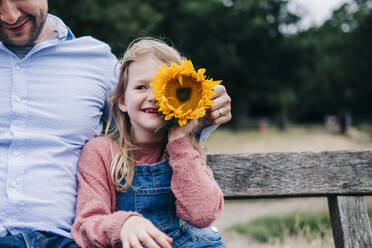 Lächelnde Tochter hält Sonnenblume, während sie mit ihrem Vater auf einer Bank sitzt - ASGF01294