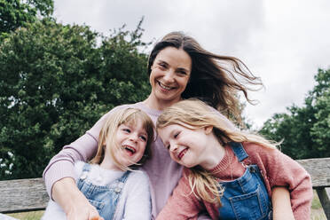 Cheerful mother with arms around daughters sitting on bench - ASGF01323