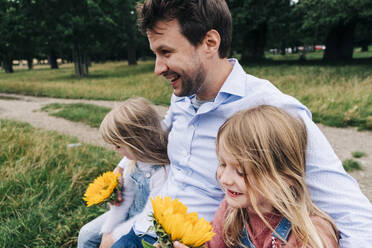 Cheerful father with arms around daughters sitting at park - ASGF01332