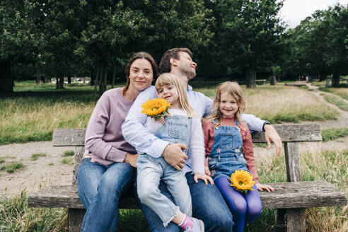 Töchter halten Sonnenblumen, während sie mit ihren Eltern auf einer Bank sitzen - ASGF01338