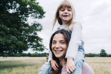 Smiling daughter sitting on woman's shoulders at meadow - ASGF01352