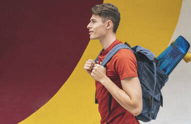 Young man carrying backpack with skateboard in front of wall - JCCMF03672