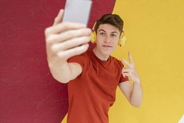 Man with headphones showing peace sign while taking selfie in front of wall - JCCMF03675