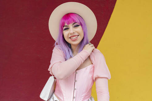 Fashionable young woman with dyed hair smiling in front of wall - JCCMF03677