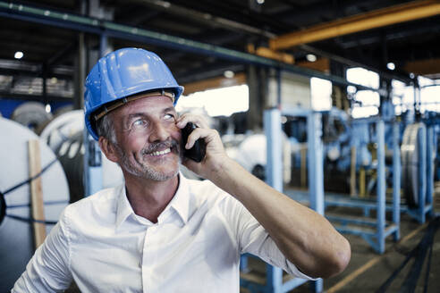 Lächelnder männlicher Ingenieur mit Schutzhelm, der in der Industrie mit einem Mobiltelefon spricht - GUSF06144