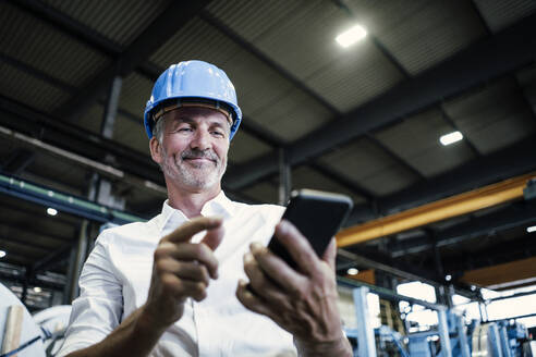 Lächelnder männlicher Ingenieur mit Schutzhelm, der in der Metallindustrie ein Mobiltelefon benutzt - GUSF06196