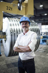 Smiling male professional with arms crossed standing in steel mill - GUSF06200
