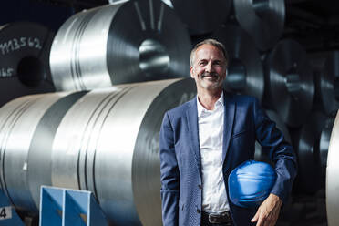 Smiling businessman holding hardhat while standing at warehouse - GUSF06215