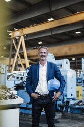 Smiling male professional holding hardhat while standing with hand in pocket at industry - GUSF06231