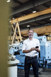 Smiling male business professional with mobile phone standing in steel mill - GUSF06234