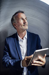 Männlicher Geschäftsmann, der ein digitales Tablet vor einer Metallblechrolle hält - GUSF06256