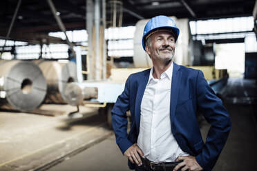 Lächelnder Geschäftsmann mit Schutzhelm, der mit der Hand auf der Hüfte im Stahlwerk steht - GUSF06282
