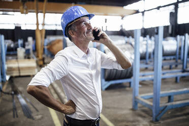 Male engineer with hardhat talking on mobile phone in warehouse - GUSF06333