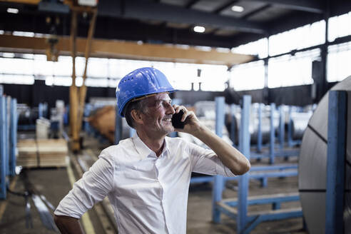 Lächelnder männlicher Ingenieur mit Schutzhelm, der in einem Stahlwerk mit einem Mobiltelefon spricht - GUSF06334