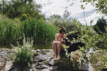 Tierhalterin streichelt Hund, während sie auf einem Felsen an einem Teich sitzt - MRRF01406