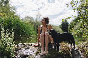 Lächelnde Frau sitzt mit Hund auf einem Felsen an einem Teich - MRRF01407