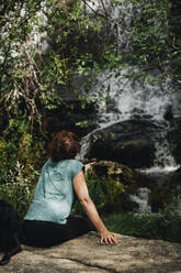 Mittlere erwachsene Frau schaut auf einen Wasserfall, während sie mit einem Hund im Wald sitzt - MRRF01433