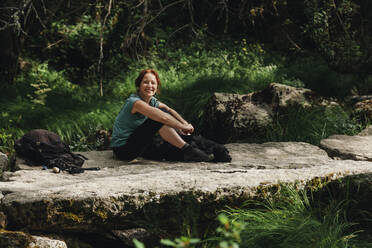 Smiling mid adult woman sitting with dog on footpath in forest - MRRF01440