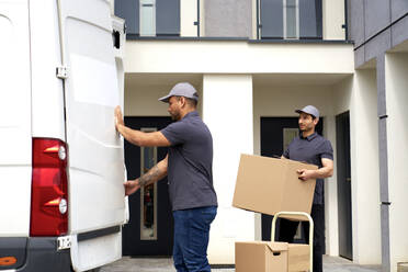 Mann, der die Tür eines Lieferwagens schließt, während sein Kollege eine Kiste vor einem Haus hält - ABIF01550