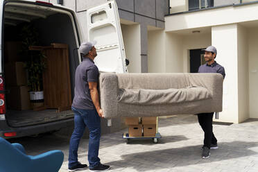 Männliche Mitarbeiter eines Lieferdienstes entladen ein Sofa aus einem Umzugswagen in der Nähe des Hauses - ABIF01554