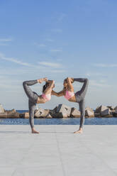 Female friends doing double dancer pose on boardwalk by sea - AFVF09100