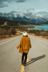 Back view of unrecognizable female tourist in casual clothes walking on David Thompson Highway near abraham lake against snowy mountains in Alberta, Canada - ADSF29231