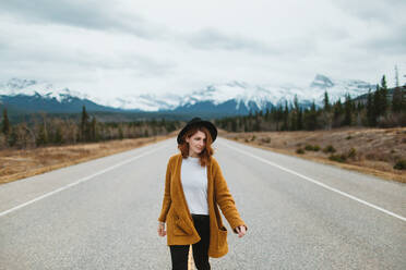 Weibliche Touristin in Freizeitkleidung geht auf dem David Thompson Highway gegen verschneite Berge in Alberta, Kanada - ADSF29234