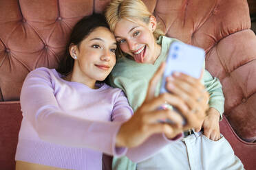 Von oben auf dem Sofa liegende, fröhliche junge Schwestern, die sich mit ihrem Handy fotografieren - ADSF29237