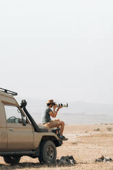 Seitenansicht eines männlichen reisenden Fotografen, der auf einem Geländewagen sitzt und ein Foto mit einer Kamera mit Teleobjektiv während einer Safari in der Savanne im Sommer macht - ADSF29249