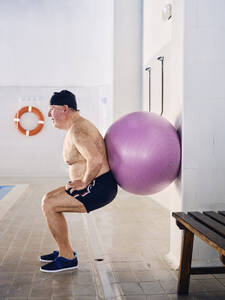 Seitenansicht eines männlichen Schwimmers mittleren Alters, der während des Aerobic-Trainings im Schwimmbad Hockübungen mit einem Fit-Ball macht - ADSF29278