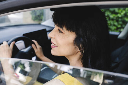 Lächelnde reife Frau, die beim Fahren eines Elektroautos vom Fenster wegschaut - JCCMF03716